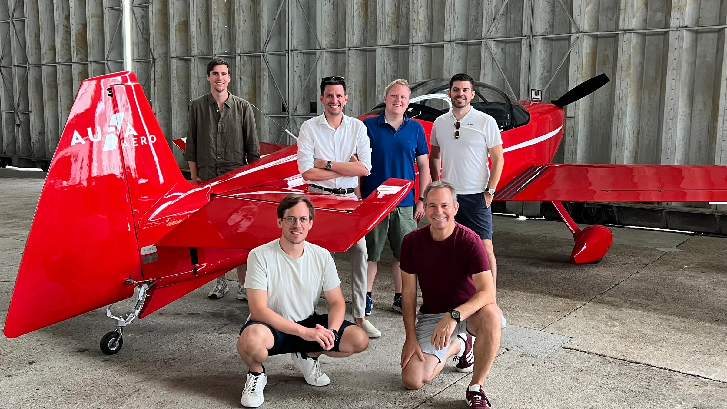 The group posing in front of a red Aura Aero INTEGRAL aircraft in a hangar